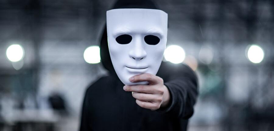 Mystery hoodie man with black mask holding white mask in his hand depciting a hacker during Halloween Cybersecurity concept