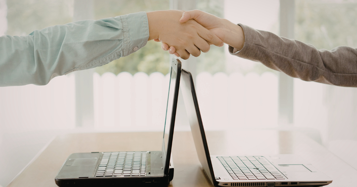 Business professionals shaking hands across two laptops, representing co-managed IT collaboration and partnership for cybersecurity and operational support.